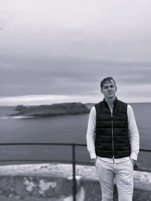 Svartvit bild på Liam Hjalmarsson som står utomhus vid ett räcke med hav och klippor i bakgrunden och under en molnig himmel.